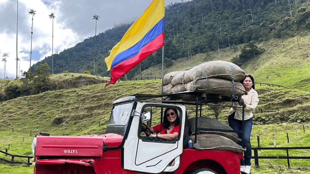 A red Willys truck with a Colombian flag, a driver, and a passenger on a grassy hillside. 3