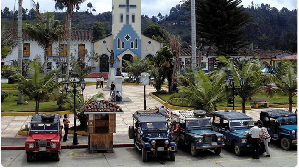 A church with a bell tower, parked vintage cars, and palm trees in a town square. 1