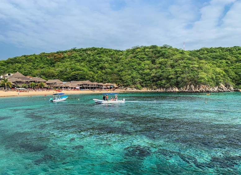 A tropical beach with clear turquoise water, boats, and thatched-roof huts. 1