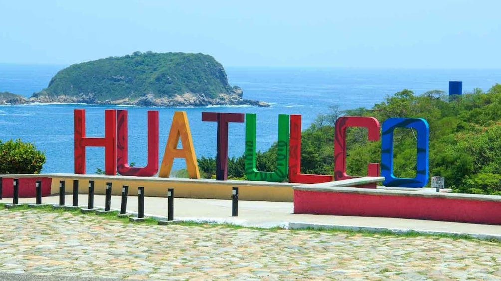 A sign with 'HUATILCO' in colorful letters, a coastal view with a small island, and a paved walkway. 4