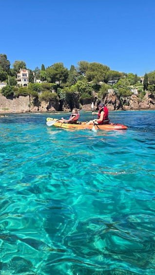 Two people kayaking on clear turquoise water with a rocky shoreline and buildings in the background. 2