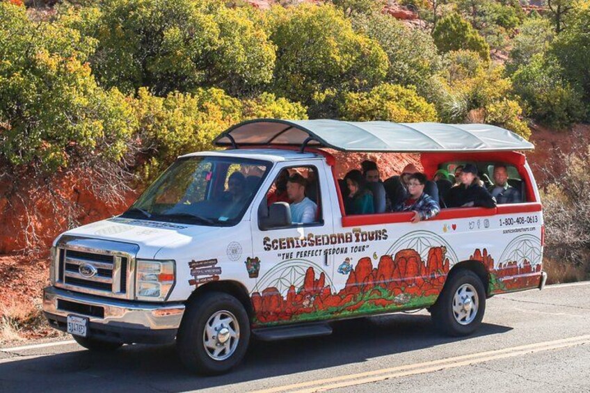 A white tour bus with red and green graphics, labeled "Scenic Sequoia Tours," driving on a road with a red rock landscape in the background. 1