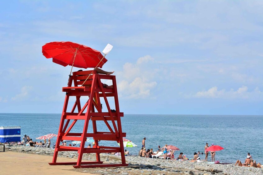 A red lifeguard tower with a red umbrella on a rocky beach with people sunbathing and swimming. 1