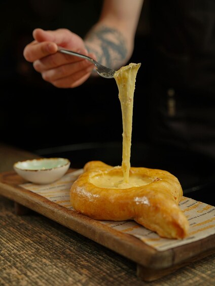 A person holding a fork with melted cheese stretching from a baked dish on a wooden board. 3