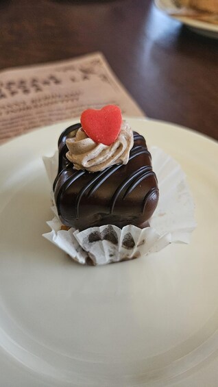 A chocolate-covered dessert with a heart-shaped decoration on top, placed on a white plate. 4