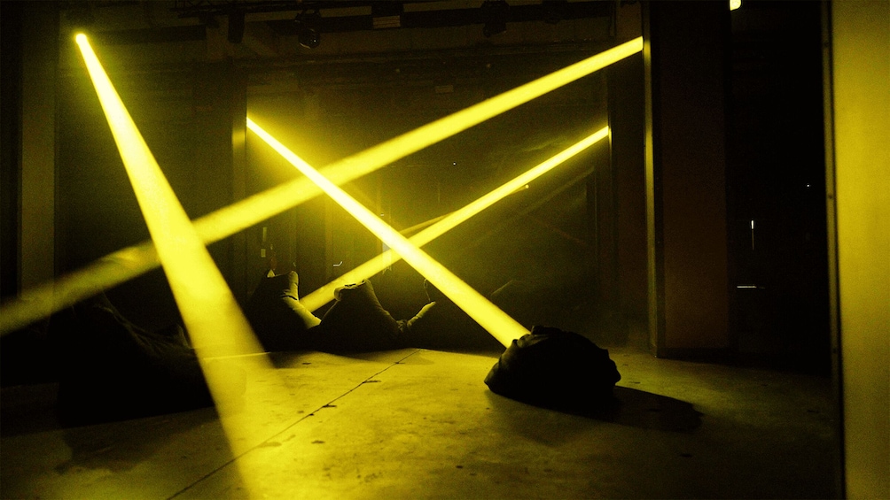 A dimly lit room with intersecting yellow light beams and a dark object on the floor. 5