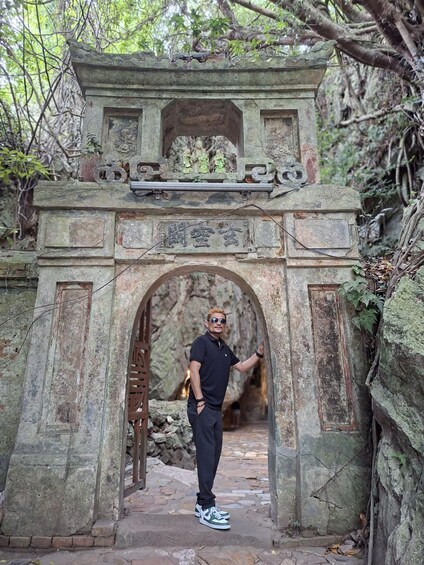 A man standing in front of an ancient stone archway with inscriptions, surrounded by greenery and a rocky path. 5