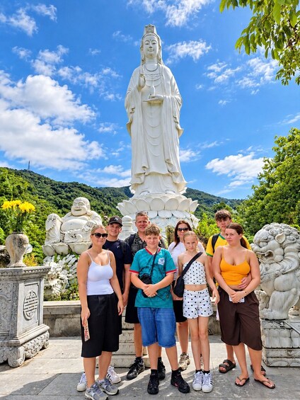 A group of people standing in front of a large white statue, with a mountain and greenery in the background. 4