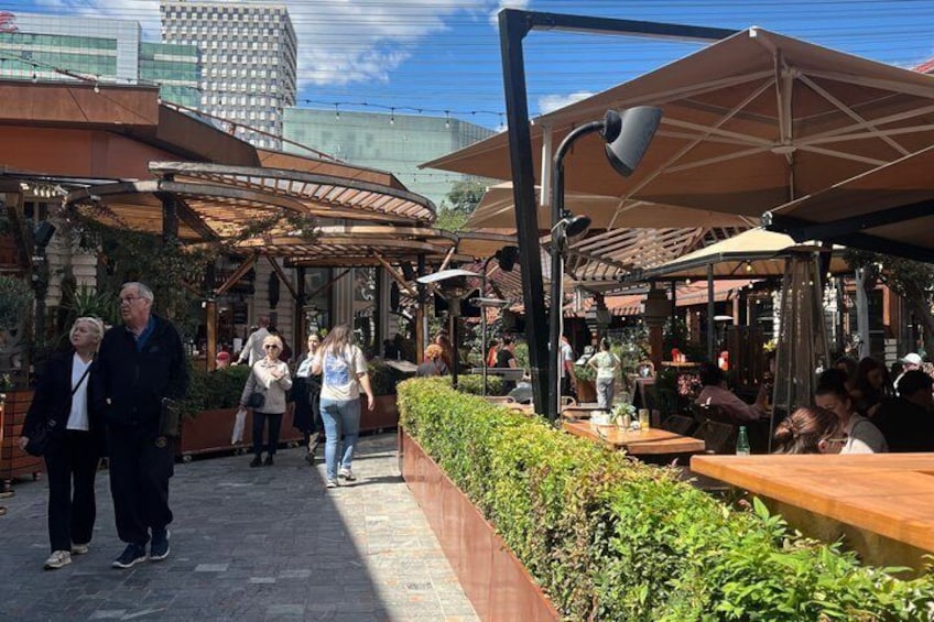 Ett utomhuskafé med träbord och parasoller, omgivet av grönska och stadens byggnader. 3