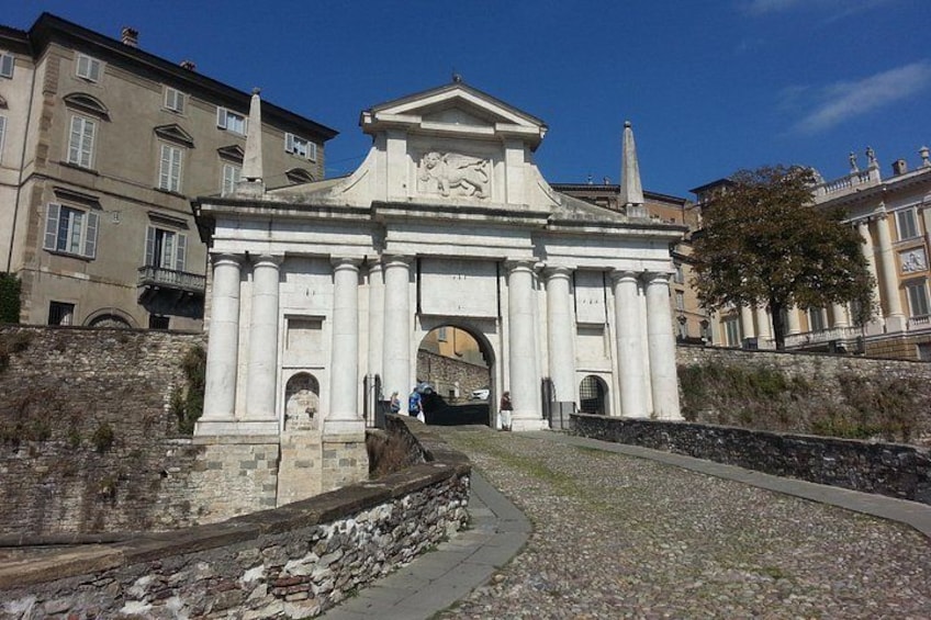 Un arco clásico con columnas, un muro de piedra y edificios al fondo. 1