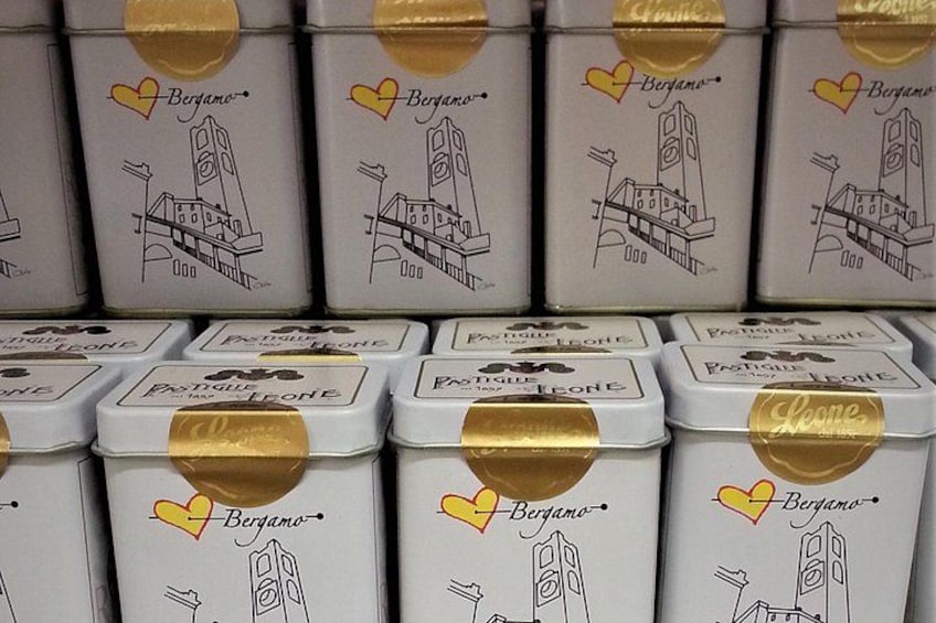 Una exhibición de cajas blancas con cintas doradas, cada una con el nombre 'Bergamo' y un boceto de un edificio. 5