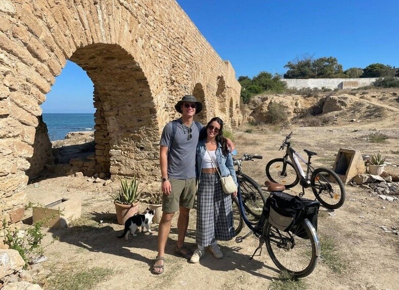 A man and a woman standing in front of an ancient stone archway with a bicycle and a cat nearby. 4