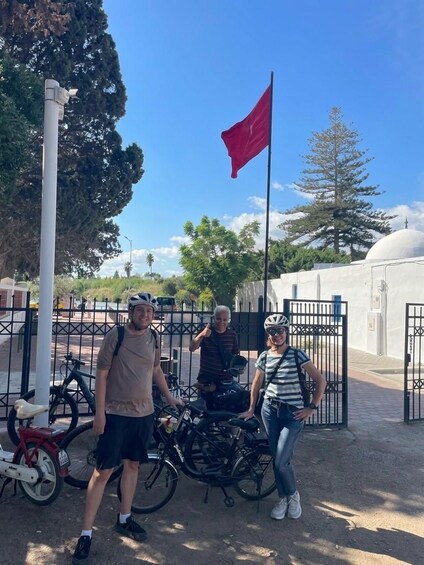 Three people standing in front of a gate with bicycles, a red flag, and a white dome structure in the background. 5