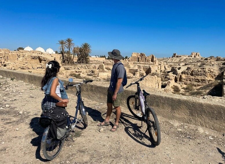A man and a woman standing by bicycles in an archaeological site with ruins and palm trees in the background. 3