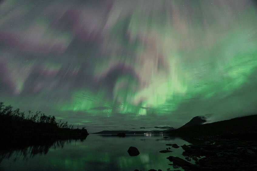 The aurora borealis over a calm lake with rocky shores and a distant mountain. 3