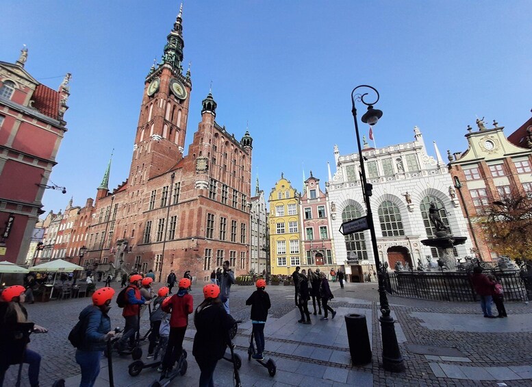 En gruppe mennesker med hjelme, måske på en guidet rundvisning, på et historisk bytorv med markante bygninger og en klar blå himmel. 5