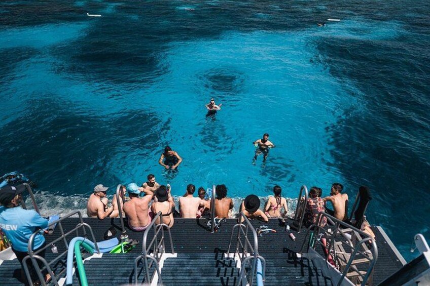 Un grupo de personas en un bote, algunas en el agua y otras en la cubierta, con un mar azul y transparente al fondo. 2