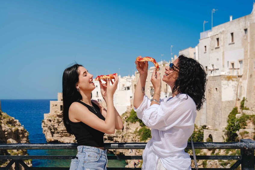 Two people holding pizza slices on a balcony with a coastal view. 1