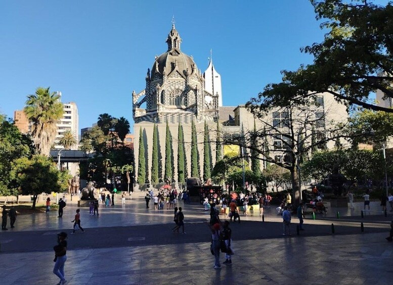 Una gran catedral amb una cúpula i agulles, envoltada d'arbres i gent en una plaça. 1