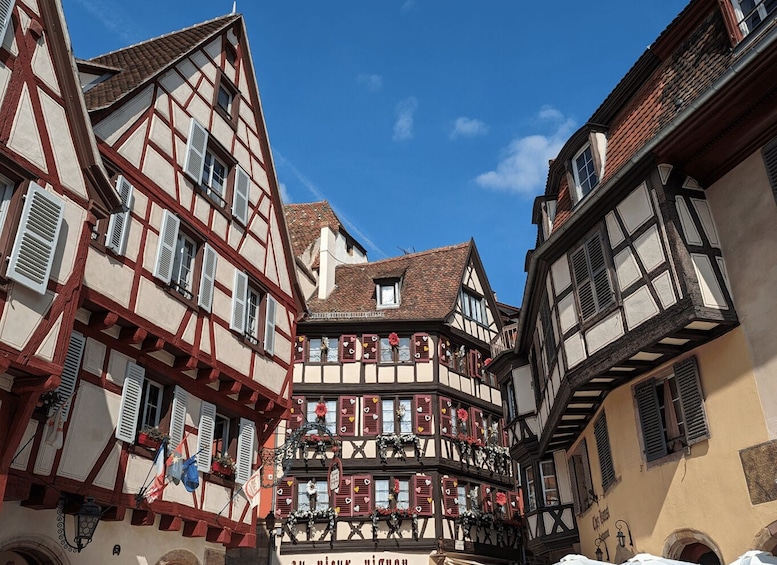 Una calle estrecha con edificios tradicionales de entramado de madera, un cielo azul claro y un letrero que dice 'Au plaisir de vivre'. 2