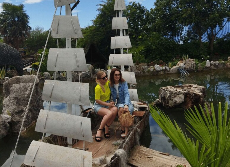 Two people sitting on a wooden dock by a calm pond with a model ship and rocks. 4