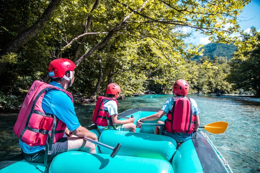 Three people in a turquoise raft with red helmets and life vests, paddling through a river surrounded by dense green foliage and hills. 2