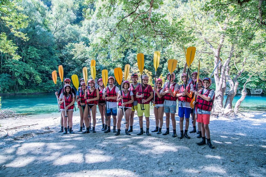 A group of people in red life jackets standing on a gravel path by a river, holding yellow paddles. 3