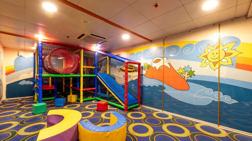 A colorful indoor play area with a climbing structure, soft play mats, and a mural with a sun and clouds. 2