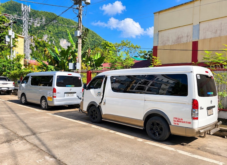Two white vans parked on a street with a mountain and greenery in the background. 5