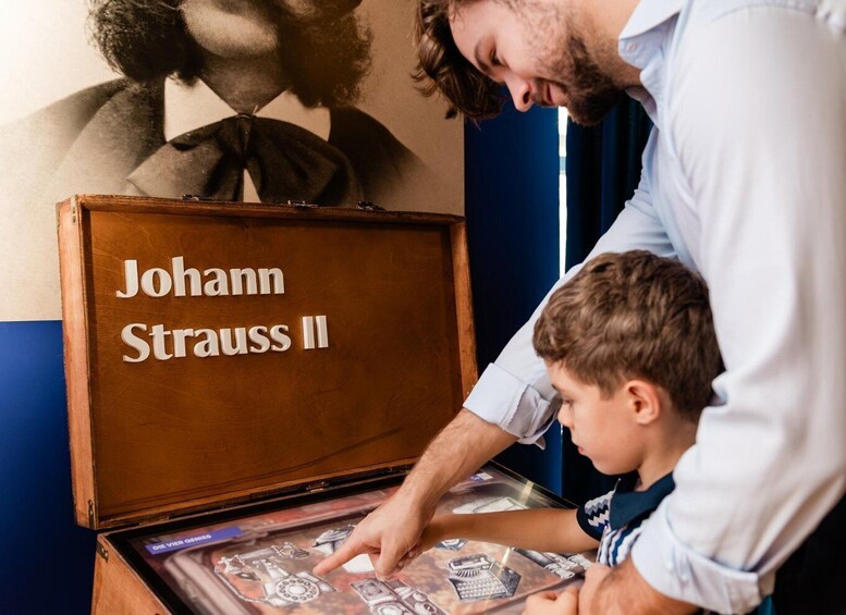 A man and a child examining a wooden box labeled 'Johann Strauss II' with sheet music inside. 2