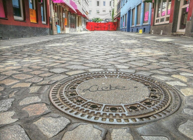 A cobblestone street with a manhole cover in the foreground and colorful buildings in the background. 5