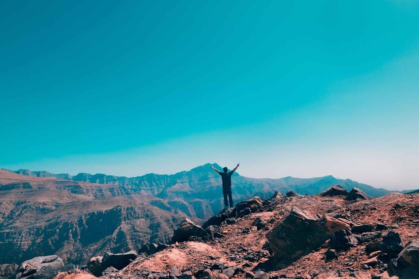 A person standing on a rocky mountain peak with arms raised, overlooking a vast mountainous landscape. 3