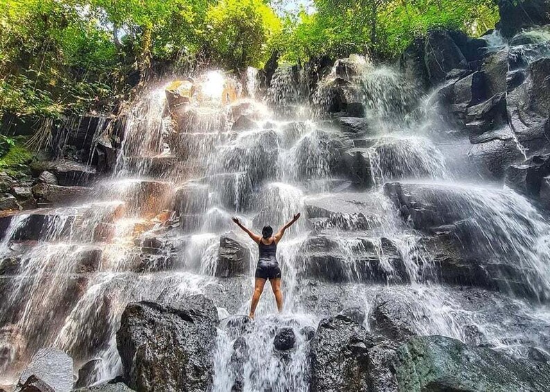 A person standing on rocks with arms raised, surrounded by a cascading waterfall and lush greenery. 5