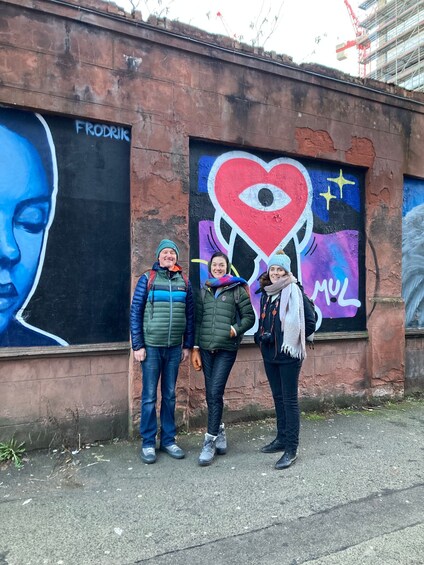 Kolme ihmistä seisoo seinän edessä, ja seinässä on graffititaidetta. 1