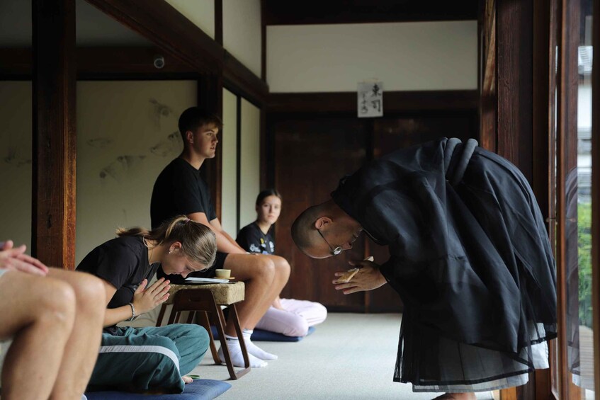 A group of people in a traditional Japanese setting, some sitting on tatami mats, others standing, and one person in black robes. 4
