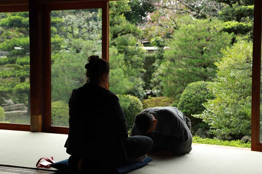 A person sitting on a mat inside a building, looking out at a garden with trees and bushes. 2