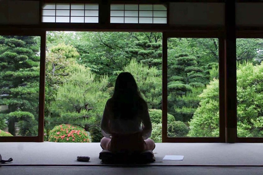 A person sitting on a tatami mat facing a large window with a view of a lush garden. 1
