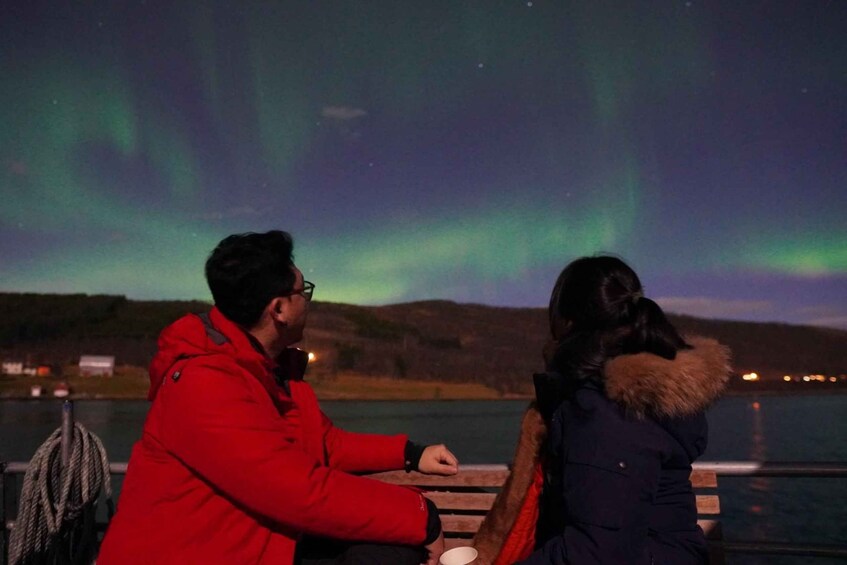 Dos personas observando las auroras boreales sobre un cuerpo de agua, con un paisaje montañoso al fondo. 4