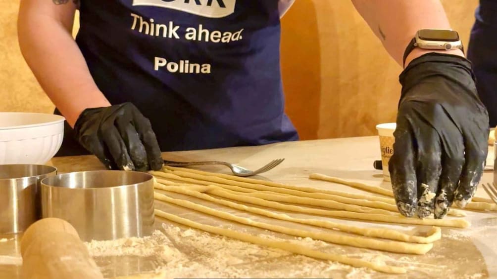 Una persona con delantal oscuro y la frase 'Think ahead Polina' preparando comida, manipulando pasta y usando un reloj en la muñeca. 5
