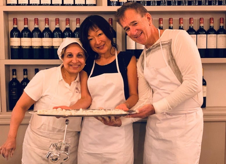 Trois personnes se trouvent dans une cuisine, portant des tabliers blancs, et on peut voir des bouteilles de vin sur des étagères à l’arrière-plan. 5