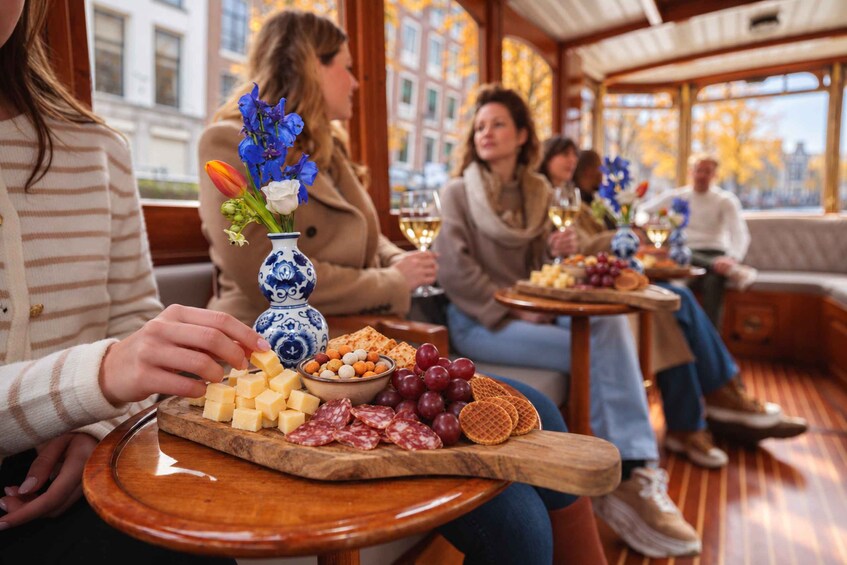 Un groupe de personnes partage un repas composé de fromage, de raisins et de fleurs, assis autour d’une table en bois dans une pièce chaleureuse et bien éclairée, dotée de grandes fenêtres. 2