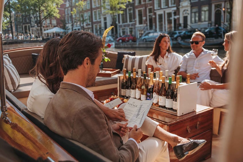 A group of people seated on a boat, enjoying a meal and drinks, with bottles of wine and a menu visible. 4