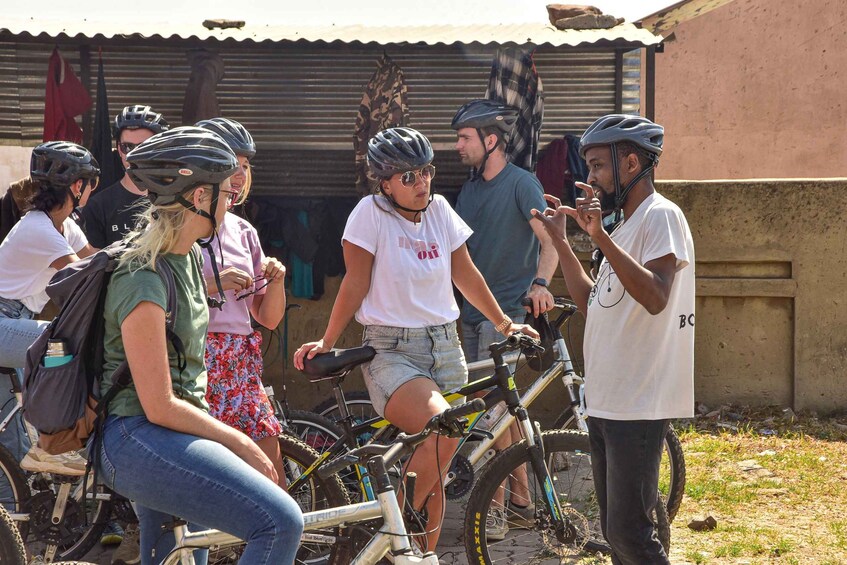 A group of cyclists gathered around a bicycle, with a corrugated metal building in the background. 1