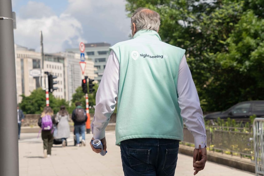 A person walking on a sidewalk wearing a light green vest with the logo 'sightseeing.lu'. 3