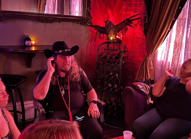 Una persona con sombrero y camisa negros sentada a una mesa con una bebida y una vela, en una habitación con iluminación roja y una escultura decorativa de un pájaro. 3