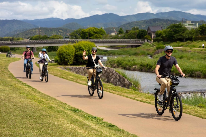 Un groupe de cyclistes sur une piste asphaltée qui longe une rivière, avec des montagnes en arrière-plan et un pont qui traverse la rivière. 1
