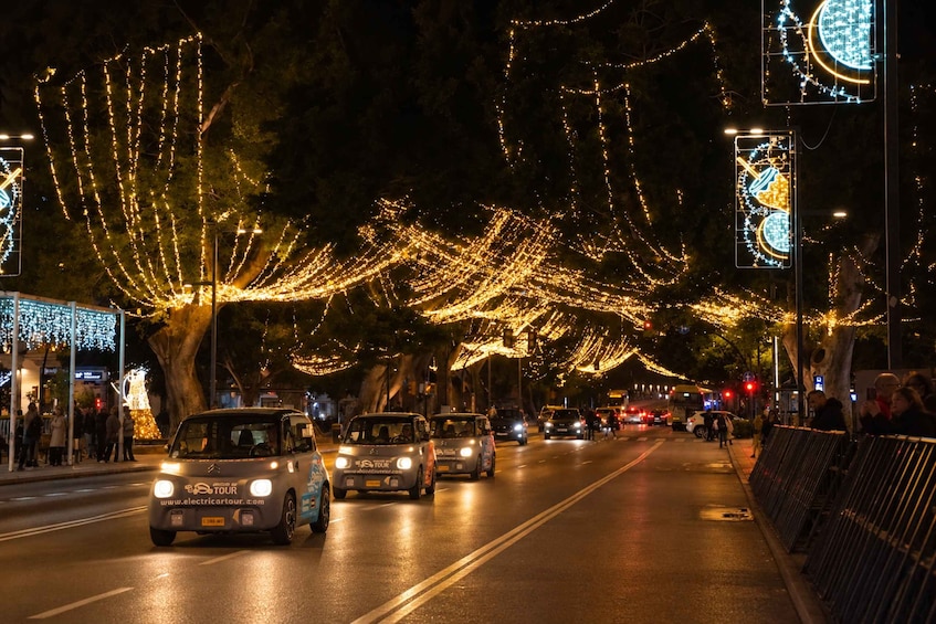 Una calle bordeada de árboles decorados con luces de adorno, vehículos circulando por la calzada y letreros iluminados. 2