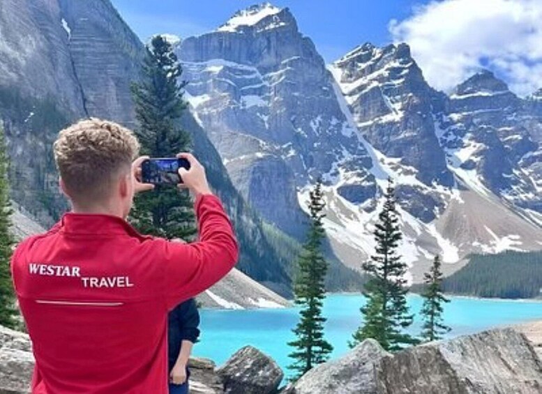 A person in a red jacket with 'WESTAR TRAVEL' on the back, taking a photo of a mountain landscape with snow-capped peaks and a turquoise lake. 2