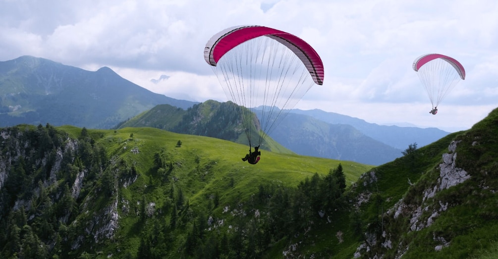 Zwei Gleitschirmflieger über einer gebirgigen Landschaft mit saftig grünen Hügeln und steinigen Klippen. 3