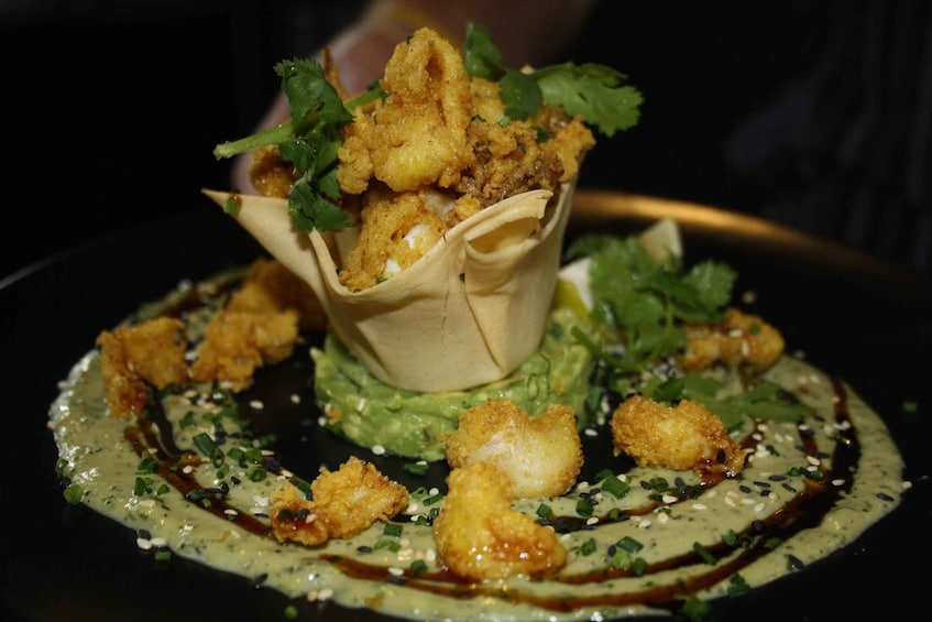 A dish with fried items in a paper cup, garnished with cilantro, and surrounded by green sauce and sesame seeds. 4